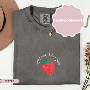 Peut inclure: T-shirt brod&eacute; de couleur poivre, taille XL-TG-EG, avec un motif de fraise et l'expression "behavior is my jam". Le t-shirt a un col rond et des manches courtes. Un collier pendentif &eacute;toile dor&eacute;e est visible.