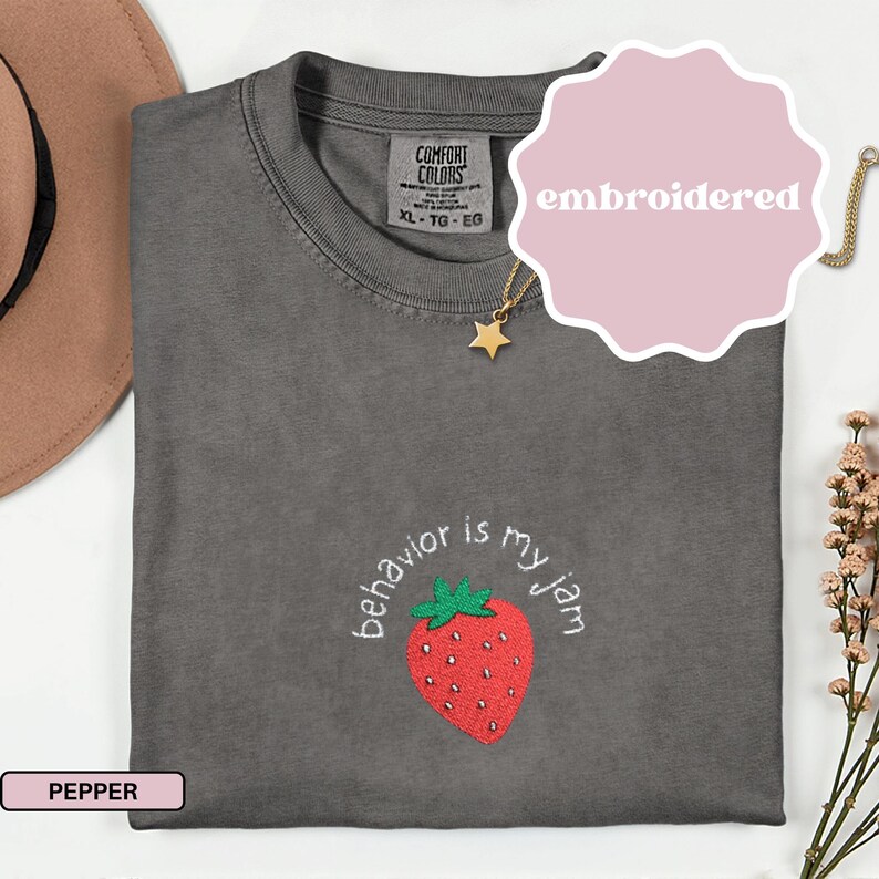 Peut inclure: T-shirt brod&eacute; de couleur poivre, taille XL-TG-EG, avec un motif de fraise et l'expression "behavior is my jam". Le t-shirt a un col rond et des manches courtes. Un collier pendentif &eacute;toile dor&eacute;e est visible.
