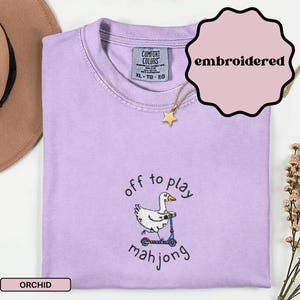 May include: A light purple, XL-sized t-shirt with the text "off to play mahjong" and an embroidered duck riding a scooter. The shirt has a Comfort Colors tag and a gold star pendant necklace. The word "embroidered" is in a pink circle.