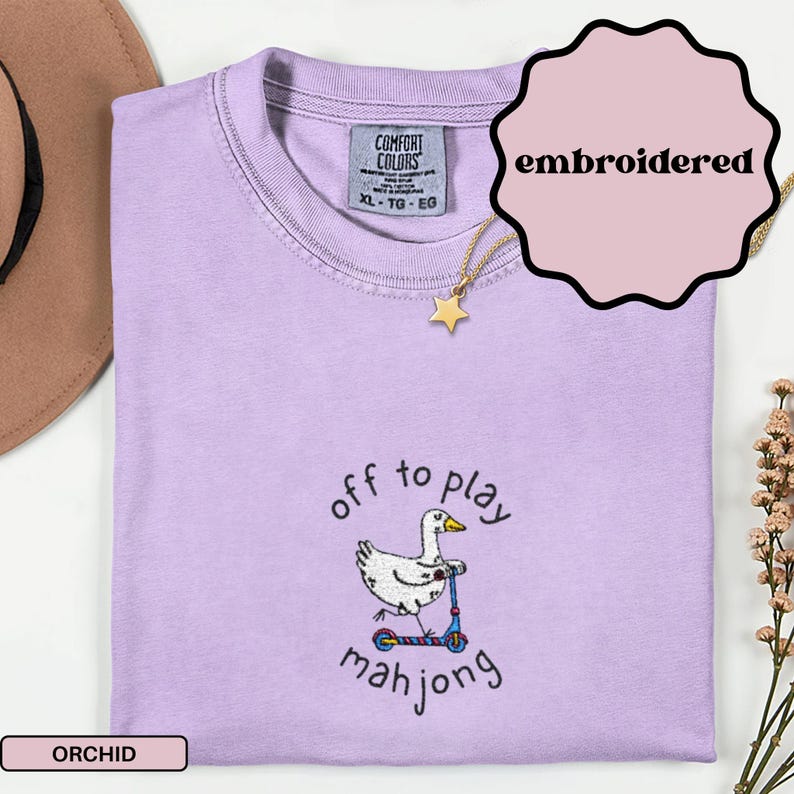 May include: A light purple, XL-sized t-shirt with the text "off to play mahjong" and an embroidered duck riding a scooter. The shirt has a Comfort Colors tag and a gold star pendant necklace. The word "embroidered" is in a pink circle.
