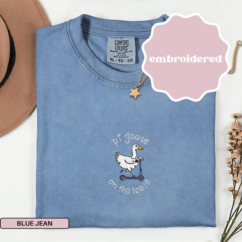 May include: A blue jean colored Comfort Colors t-shirt with an embroidered design. The design features a white goose riding a scooter with the text "pt goose on the loose". The shirt is size XL.