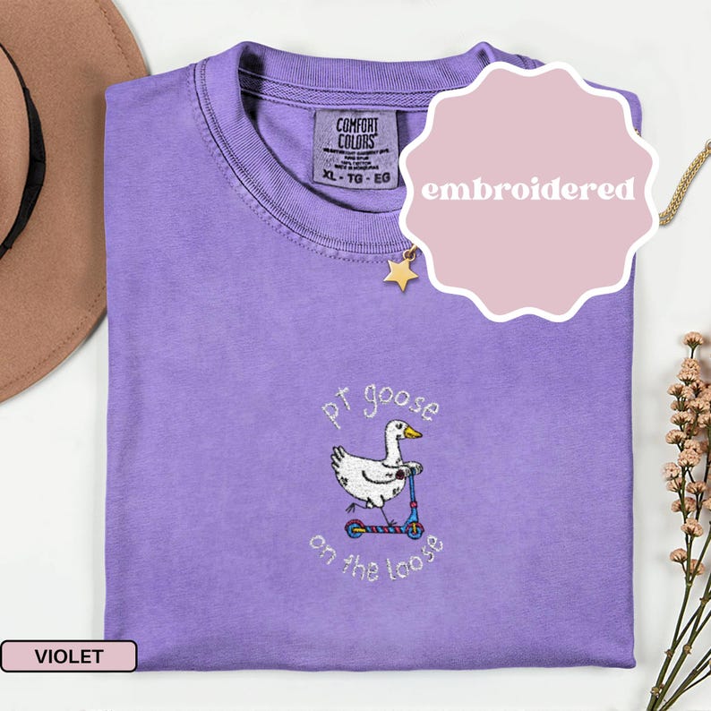 May include: A violet Comfort Colors t-shirt with an embroidered design. The design features a white goose riding a blue and red scooter, with the text "pt goose on the loose". The shirt has a crew neck and a tag that says "XL - TG - EG".