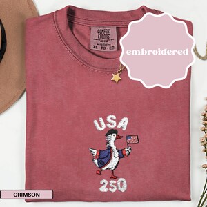 May include: Crimson-colored t-shirt with an embroidered design featuring a goose wearing a hat and jacket, holding a small American flag. The word "USA" is above the goose, and "250" is below. The shirt has a tag that says "Comfort Colors".