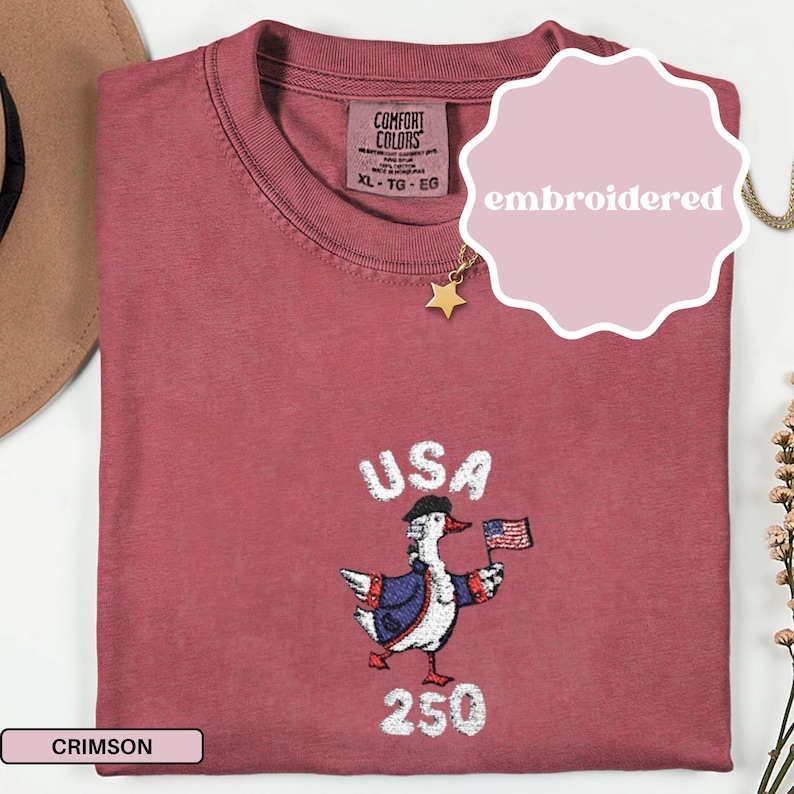 May include: Crimson-colored t-shirt with an embroidered design featuring a goose wearing a hat and jacket, holding a small American flag. The word "USA" is above the goose, and "250" is below. The shirt has a tag that says "Comfort Colors".