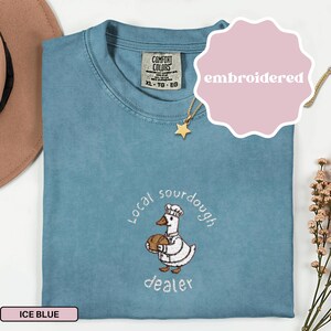 Puede incluir: Camiseta azul hielo con un dise&ntilde;o bordado. El dise&ntilde;o presenta un ganso de dibujos animados con gorro y delantal de chef, sosteniendo un pan, con las palabras "Local sourdough dealer" alrededor. Se ve un collar de estrella dorada.