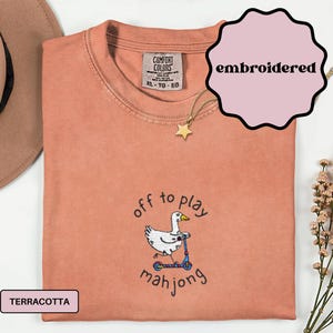 May include: Terracotta-colored, embroidered t-shirt with a cartoon duck riding a scooter, and the words "off to play mahjong". The shirt has a Comfort Colors tag and a gold star necklace. The shirt is XL.
