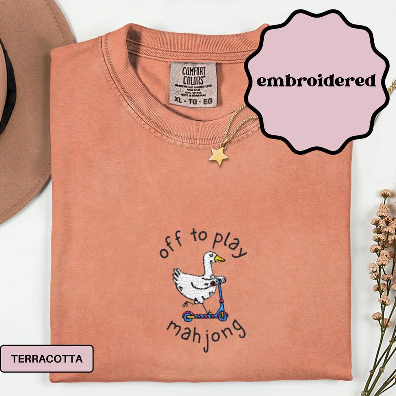May include: Terracotta-colored, embroidered t-shirt with a cartoon duck riding a scooter, and the words "off to play mahjong". The shirt has a Comfort Colors tag and a gold star necklace. The shirt is XL.