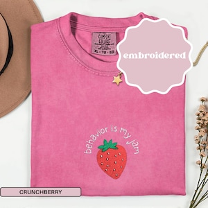 Peut inclure: Un t-shirt rose Comfort Colors avec le texte "behavior is my jam" et une fraise brod&eacute;e. Le t-shirt a un col rond et des manches courtes. Le mot "embroidered" est sur un autocollant rose. La couleur est "Crunchberry".