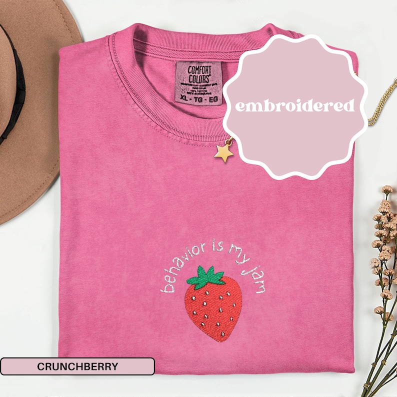 Peut inclure: Un t-shirt rose Comfort Colors avec le texte "behavior is my jam" et une fraise brod&eacute;e. Le t-shirt a un col rond et des manches courtes. Le mot "embroidered" est sur un autocollant rose. La couleur est "Crunchberry".