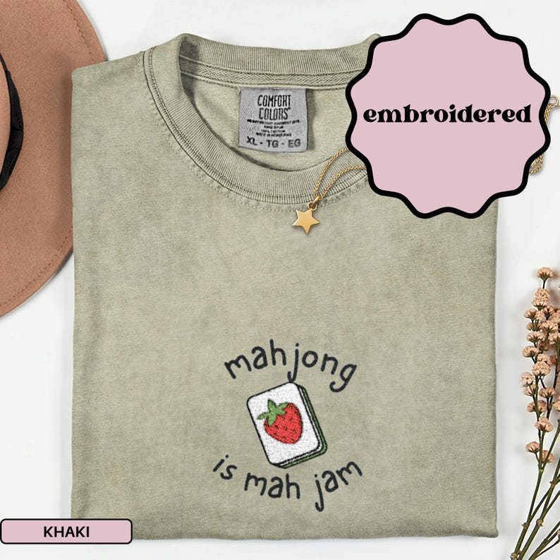 May include: Khaki-colored, embroidered t-shirt with the text "mahjong is mah jam" and a strawberry tile design. The shirt has a Comfort Colors tag and a gold star necklace. The word "embroidered" is in a pink bubble.
