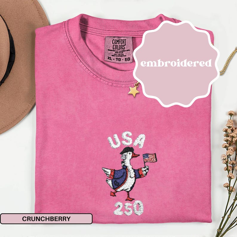 May include: A Crunchberry pink t-shirt with an embroidered design featuring a duck wearing a patriotic outfit and holding a US flag. The word "USA" is above the duck, and "250" is below.