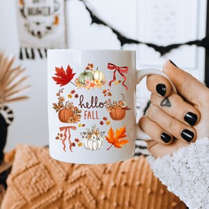 May include: White ceramic mug with a fall-themed design. The mug features pumpkins, leaves, and flowers in shades of orange, red, and yellow. The text "hello fall" is printed on the mug.
