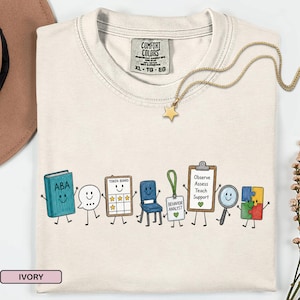 Op de afbeelding: Een ivoorkleurig t-shirt met een afbeelding van stripfiguren, waaronder een boek met het label ABA, een tekstballon, een tokenbord, een stoel, een badge voor gedragsanalist, een klembord met de tekst Observe Assess Teach Support, een vergrootglas.