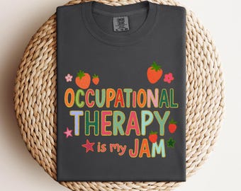 Op de afbeelding: Donkergrijs t-shirt met de tekst "OCCUPATIONAL THERAPY is my JAM" in kleurrijke letters, versierd met aardbeien en bloemen. Het shirt wordt gepresenteerd op een gevlochten mand.