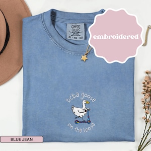 Peut inclure: T-shirt bleu jean Comfort Colors avec un motif brodé. Le motif représente une oie sur un scooter avec le texte "bcba goose on the loose". Le t-shirt est de taille XL-TG-EG.