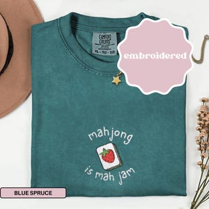May include: Blue spruce t-shirt with an embroidered mahjong tile design. The tile features a red strawberry and the text "mahjong is mah jam". The shirt is a Comfort Colors XL-TG-EG.