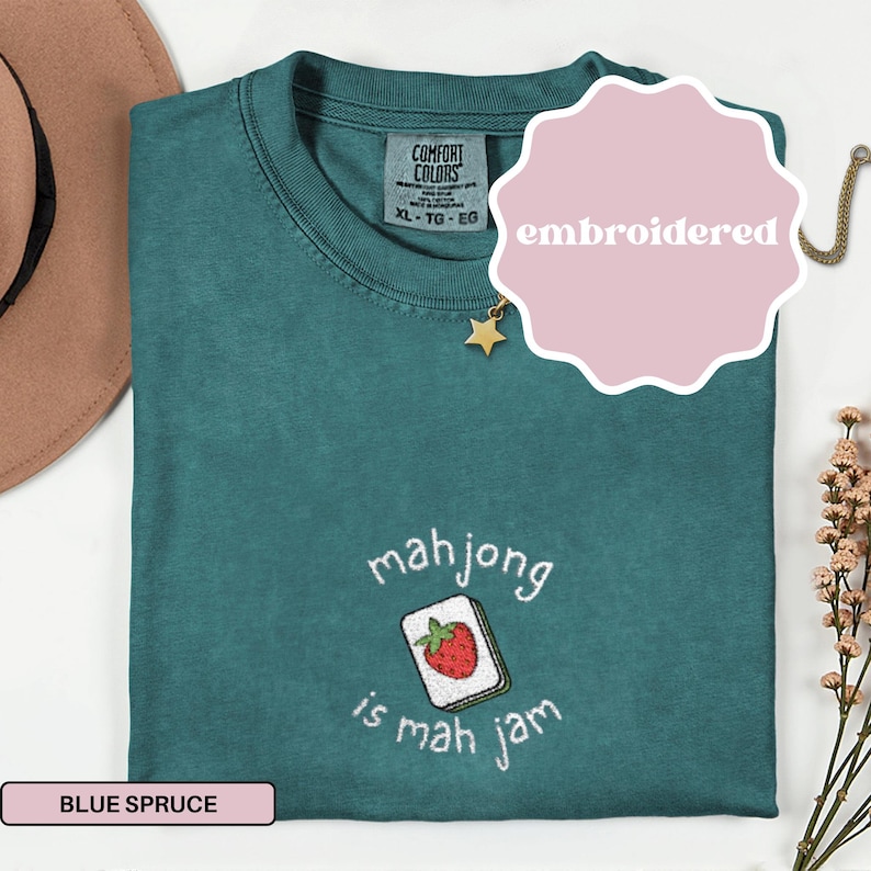 May include: Blue spruce t-shirt with an embroidered mahjong tile design. The tile features a red strawberry and the text "mahjong is mah jam". The shirt is a Comfort Colors XL-TG-EG.