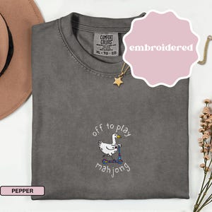 May include: Pepper-colored Comfort Colors t-shirt with an embroidered design. The design features a white goose riding a scooter with the text "off to play mahjong". A pink label says "embroidered". A gold star necklace is visible.