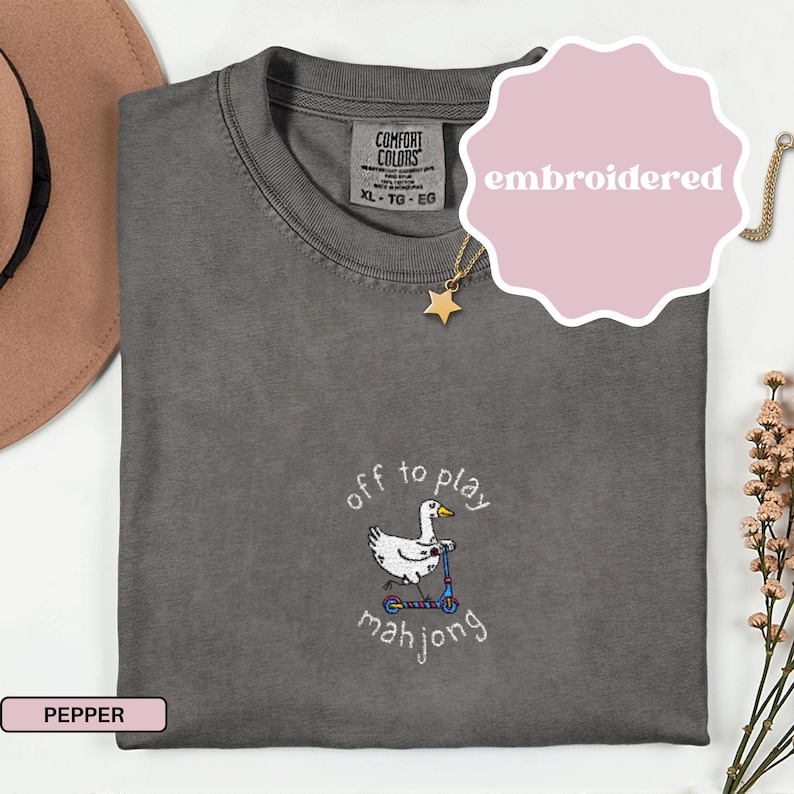May include: Pepper-colored Comfort Colors t-shirt with an embroidered design. The design features a white goose riding a scooter with the text "off to play mahjong". A pink label says "embroidered". A gold star necklace is visible.