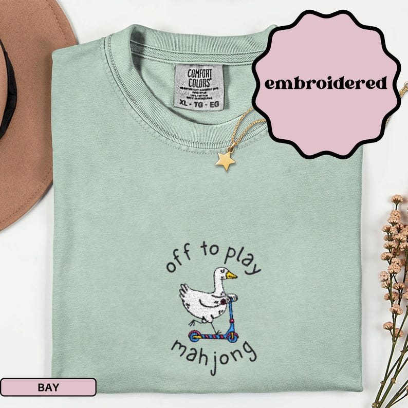 May include: A light green Comfort Colors t-shirt with an embroidered design. The design features a white duck riding a blue scooter, with the words "off to play mahjong" in black script. A gold star pendant hangs from the neckline.