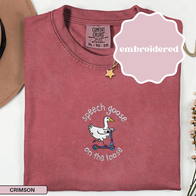 May include: Crimson-colored embroidered t-shirt with a crew neck. The shirt features an embroidered design of a white goose riding a blue scooter, with the text "speech goose on the loose". The shirt has a Comfort Colors tag.