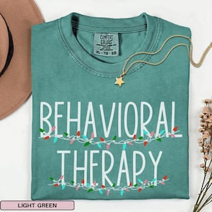 Op de afbeelding: Een blauwgroen T-shirt met ronde hals en een Comfort Colors-label aan de binnenkant van de halslijn. Het shirt heeft de witte tekst BEHAVIORAL THERAPY in een schreefloos lettertype, geaccentueerd door illustraties van kleurrijke kerstverlichting.