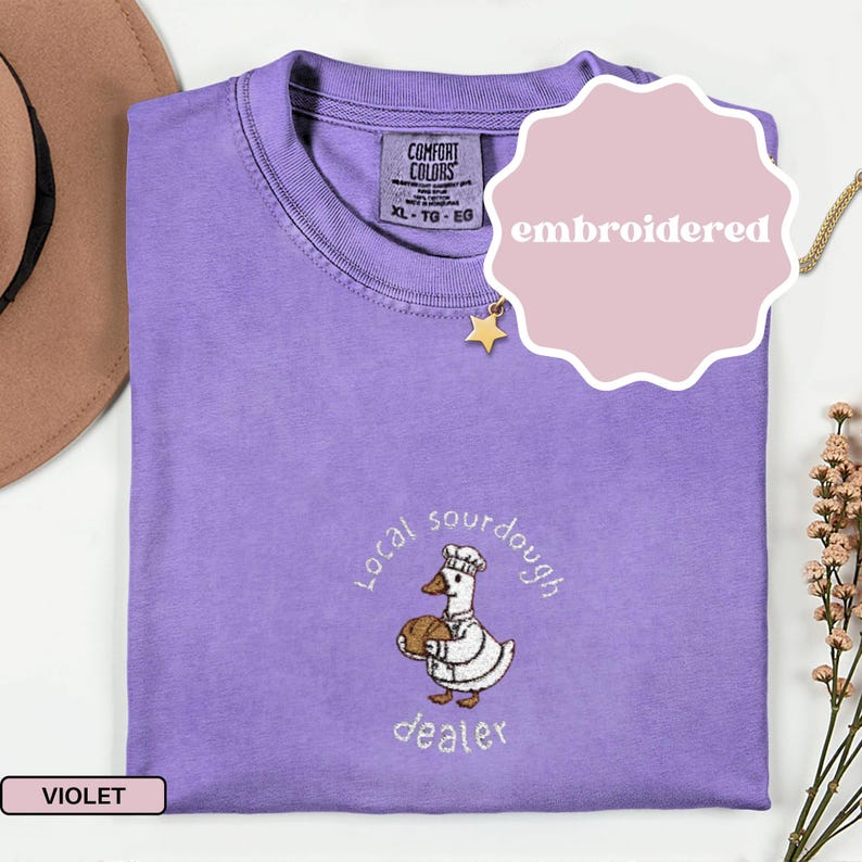 Puede incluir: Camiseta violeta Comfort Colors con un dise&ntilde;o bordado de un ganso sosteniendo pan, con las palabras "Local sourdough dealer". La camiseta tiene cuello redondo y una etiqueta que dice XL-TG-EG.