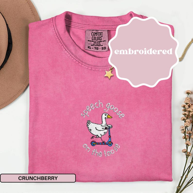 May include: A pink Comfort Colors t-shirt with an embroidered design. The design features a white goose riding a blue scooter, with the words "speech goose on the loose". The shirt is labeled "Crunchberry" and is size XL-TG-EG.