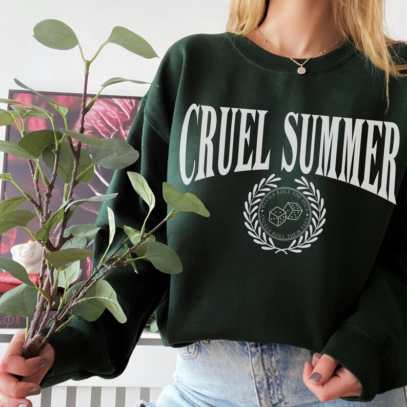 May include: A dark green sweatshirt with the text "CRUEL SUMMER" in white letters. The sweatshirt has a white graphic of a laurel wreath with dice inside and the text "DEVILS ROLL THE DICE, ANGELS ROLL THEIR EYES".