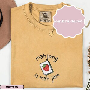 May include: Mustard-colored Comfort Colors t-shirt with embroidered text and graphic. The graphic says "mahjong is mah jam" with a strawberry tile. The shirt is size XL.