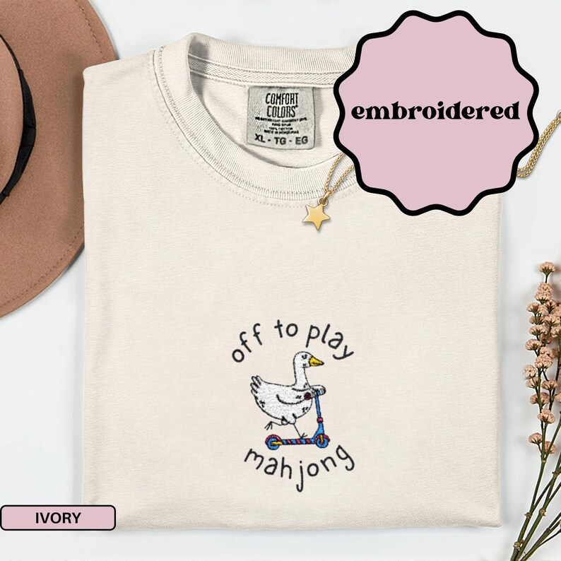 May include: Ivory-colored t-shirt with an embroidered design of a white goose riding a blue scooter, with the text "off to play mahjong". The shirt has a Comfort Colors tag and a gold necklace.