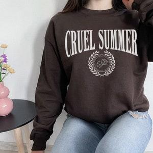 May include: A brown sweatshirt with the text "CRUEL SUMMER" in white lettering. The sweatshirt has a white graphic of a laurel wreath with dice inside, and the text "ROLL THE DICE" above the wreath.