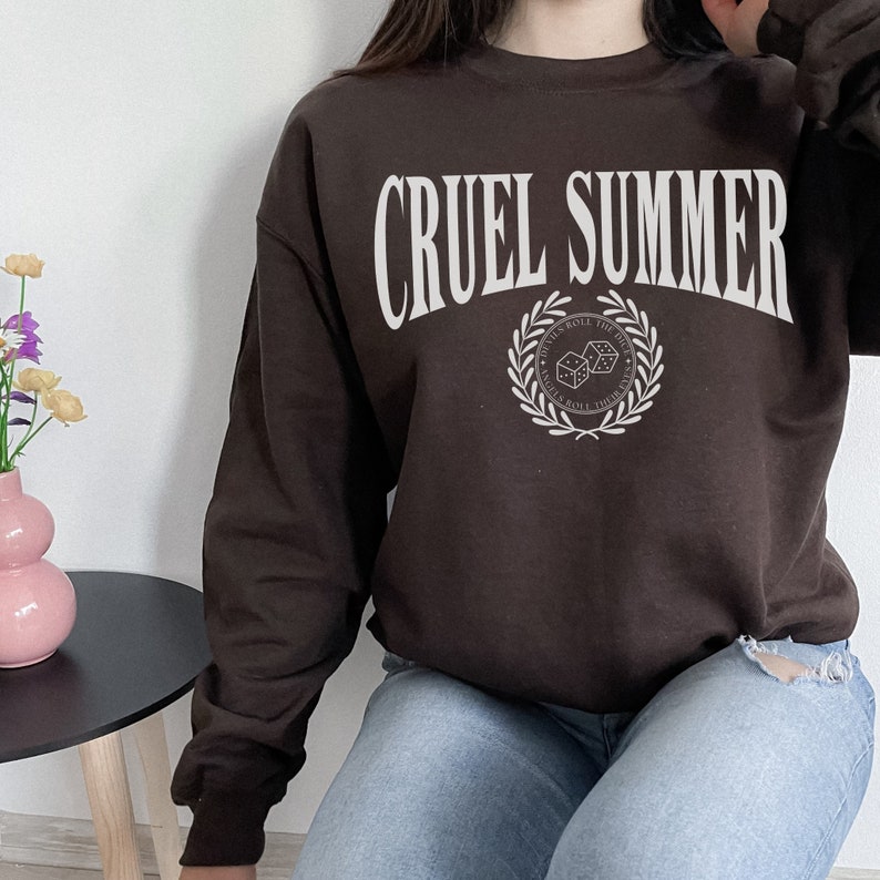 May include: A brown sweatshirt with the text "CRUEL SUMMER" in white lettering. The sweatshirt has a white graphic of a laurel wreath with dice inside, and the text "ROLL THE DICE" above the wreath.