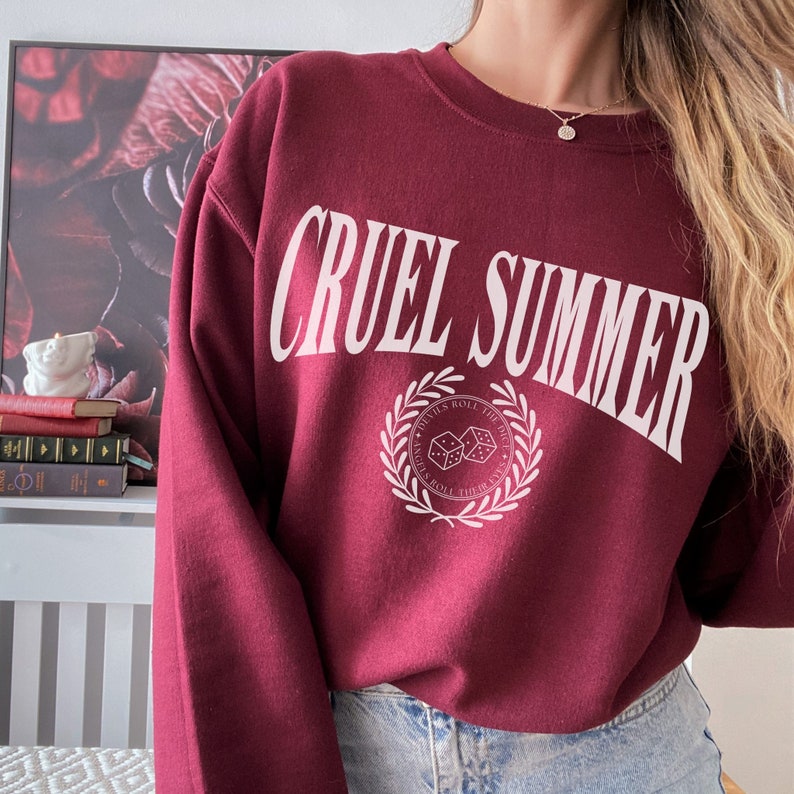 May include: Burgundy sweatshirt with white text that reads "Cruel Summer" and a white graphic of dice with a laurel wreath around them. The text around the dice reads "Devils Roll The Dice, Nights Roll Their Lives."