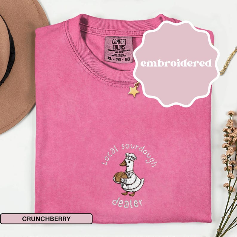 Puede incluir: Camiseta rosa con un dise&ntilde;o bordado de un pato con gorro de chef y pan. El texto "Local sourdough dealer" est&aacute; bordado encima y debajo del pato. Tambi&eacute;n se ve la palabra "Crunchberry".