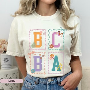 Pode incluir: Camiseta cor marfim com as letras B, C, B e A em desenhos coloridos com molduras florais. A camiseta tem gola redonda e mangas curtas. A palavra "IVORY" está impressa na parte inferior da imagem.