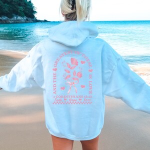 May include: A light blue hoodie with a pink graphic design that says "And the greatest of these is love" with a flower and hearts. The graphic also includes the text "1 Corinthians 13:13" and "Faith, Hope, Love".