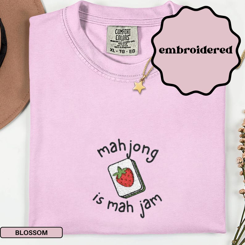 May include: A light pink t-shirt with the text "mahjong is mah jam" and an embroidered mahjong tile with a strawberry. The shirt has a crew neck and a Comfort Colors tag. A gold star necklace is visible.