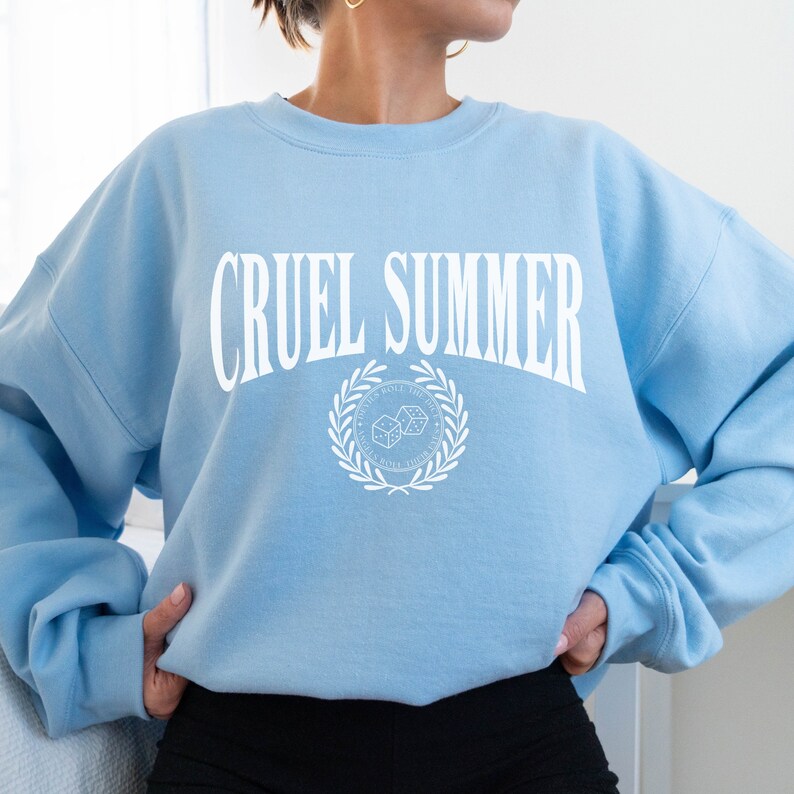 May include: Light blue sweatshirt with the text "CRUEL SUMMER" in white, featuring a white graphic of two dice with a laurel wreath around them.