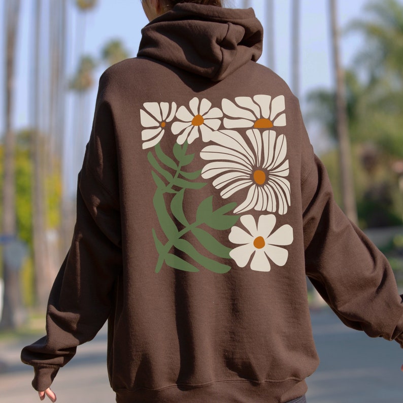 Puede incluir: Una sudadera con capucha marr&oacute;n con un estampado floral de margaritas blancas en la espalda. El estampado presenta un grupo de margaritas con hojas verdes.