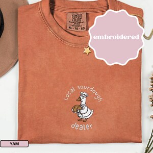 Puede incluir: Camiseta naranja quemado con un dise&ntilde;o bordado de un ganso con gorro de chef y una hogaza de pan. El texto "Local sourdough dealer" est&aacute; bordado debajo. La camiseta tiene una etiqueta Comfort Colors y est&aacute; bordada.