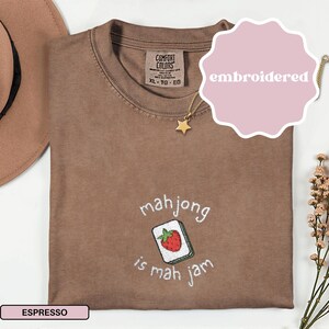 May include: Espresso-colored t-shirt with the embroidered phrase "mahjong is mah jam" and a strawberry tile design. The shirt has a crew neck and short sleeves. A gold star pendant necklace is draped on the shirt.