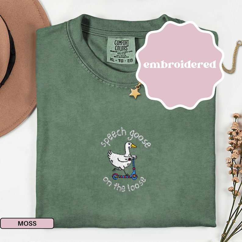 May include: Moss green t-shirt with an embroidered design of a goose on a scooter, with the text "speech goose on the loose". The shirt has a crew neck and short sleeves. The tag reads "Comfort Colors".