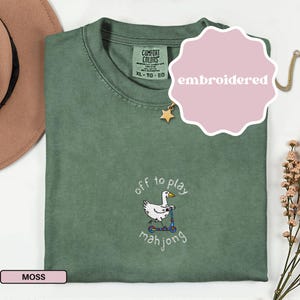 May include: A moss green t-shirt with an embroidered design. The design features a white duck riding a scooter with the words "off to play mahjong". The shirt has a crew neck and short sleeves. The word "embroidered" is on a pink sticker.