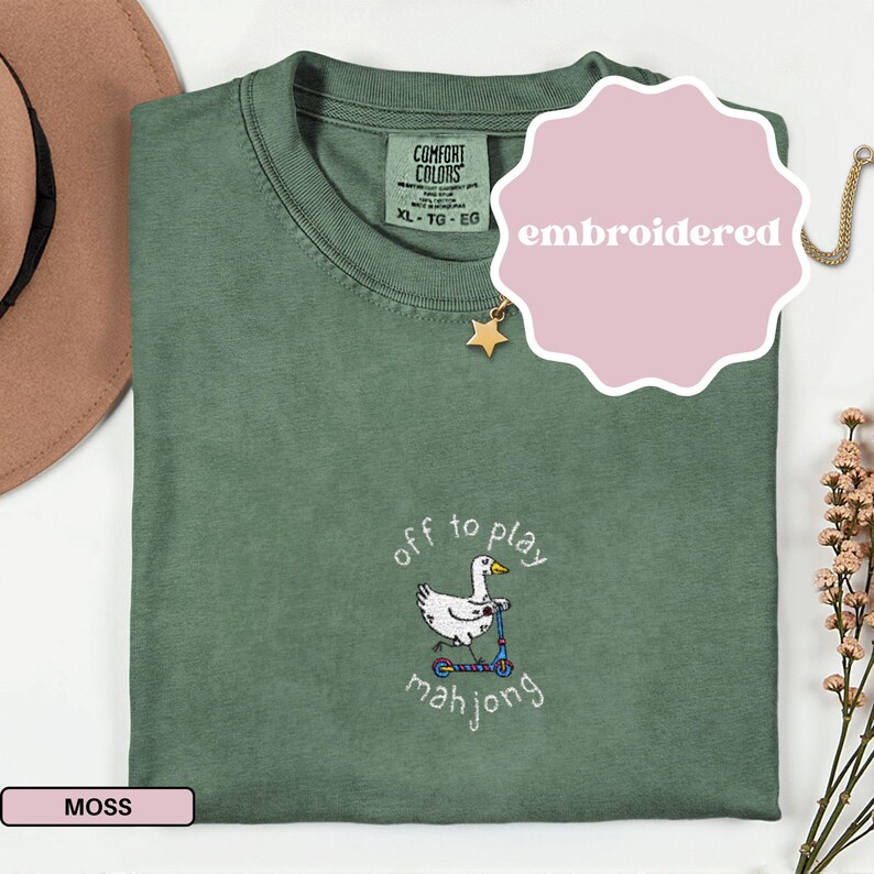 May include: A moss green t-shirt with an embroidered design. The design features a white duck riding a scooter with the words "off to play mahjong". The shirt has a crew neck and short sleeves. The word "embroidered" is on a pink sticker.