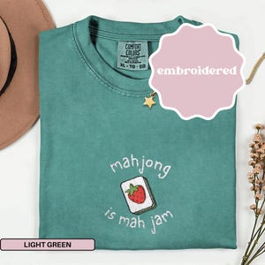 May include: Light green t-shirt with embroidered text and a strawberry mahjong tile design. The text reads "mahjong is mah jam". The shirt has a Comfort Colors tag and a pink "embroidered" tag.