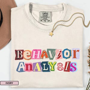 Puede incluir: Camiseta de color marfil con las palabras "BEHAVIOR ANALYSIS" en un diseño de letras recortadas multicolores. Un collar con colgante de estrella dorada descansa sobre la camiseta. La camiseta tiene una etiqueta Comfort Colors.