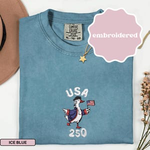 May include: Ice blue t-shirt with an embroidered design featuring a patriotic duck holding a US flag. The shirt has the word "USA" above the duck and the number "250" below. The shirt is labeled "XL - TG - EG".