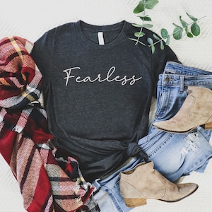 May include: A dark gray t-shirt with the word "Fearless" written in a white script font. The shirt is laid out on a white surface with a plaid scarf, blue jeans, and brown ankle boots.