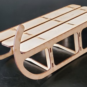 May include: A small wooden sled with a natural finish. The sled has curved runners and a slatted seat. The sled is made of light-coloured wood and appears to be a decorative item or a small toy. The sled is approximately 30 cm long.